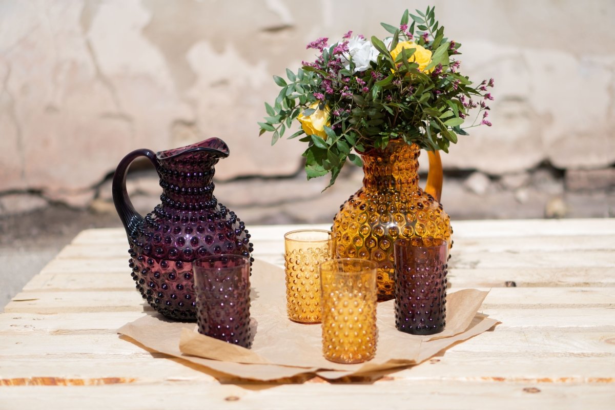 Violet Hobnail Tumblers on a wooden pallet and a light brown fabric, surrounded by products from the Hobnail collection with a broken wall in the background