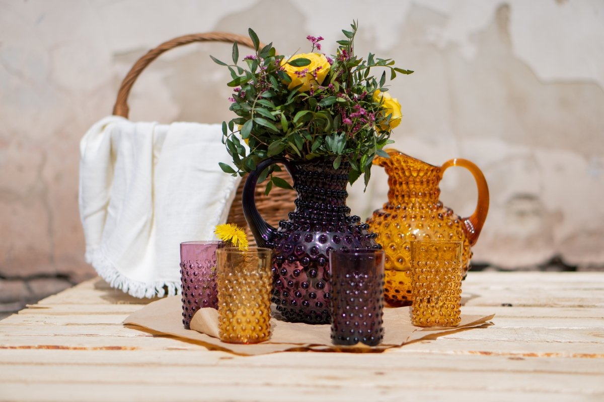 Violet Hobnail Jug on a wooden table with purple and yellow flowers, surrounded by Hobnail collection products and a wooden bag and a broken wall in the background