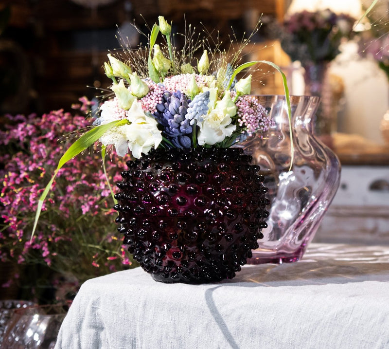 Violet Hobnail Vase on a white fabric with vase in the background with colorful flowers