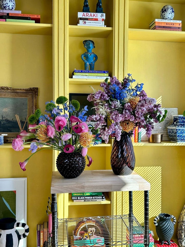 Violet Marika Cone Vase in a bookcase, on a piece of wooden furniture and with a colourful bouquet of flowers 