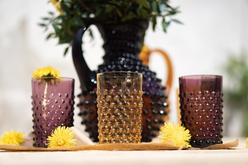 Violet Hobnail Tumblers on a wooden palette and a light brown fabric, surrounded by Hobnail collection products and yellow flowers